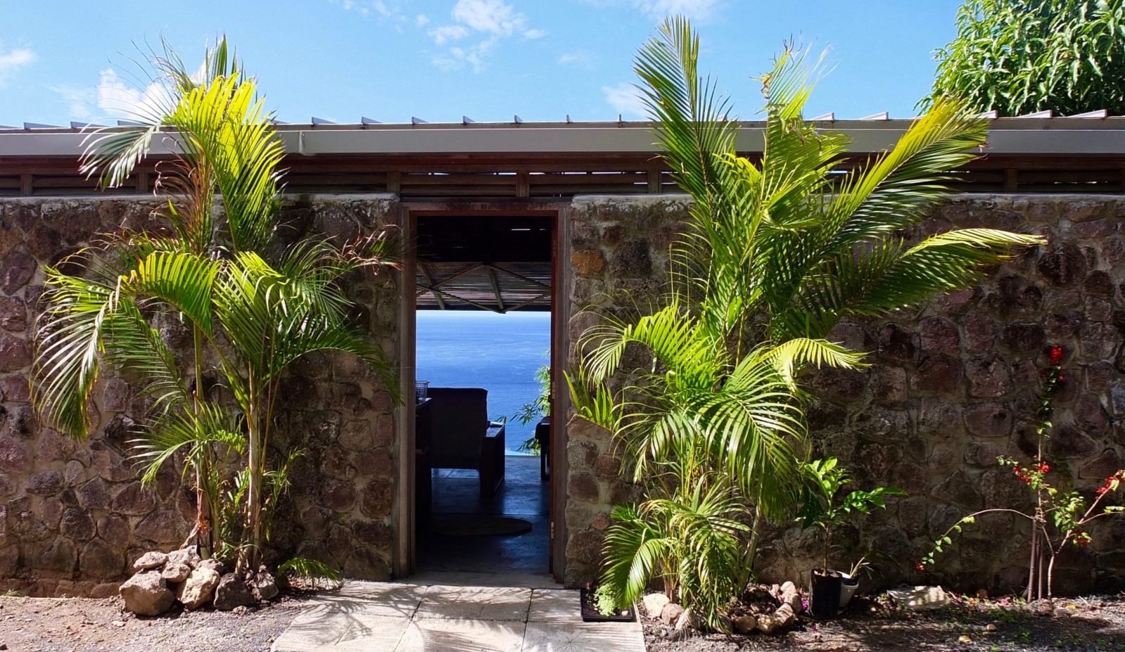 The front door of the Lodge at Cosmos St Lucia sits within a stone wall surrounded by palm fronds. Through the doorway the blue of the Caribbea Sea is visible