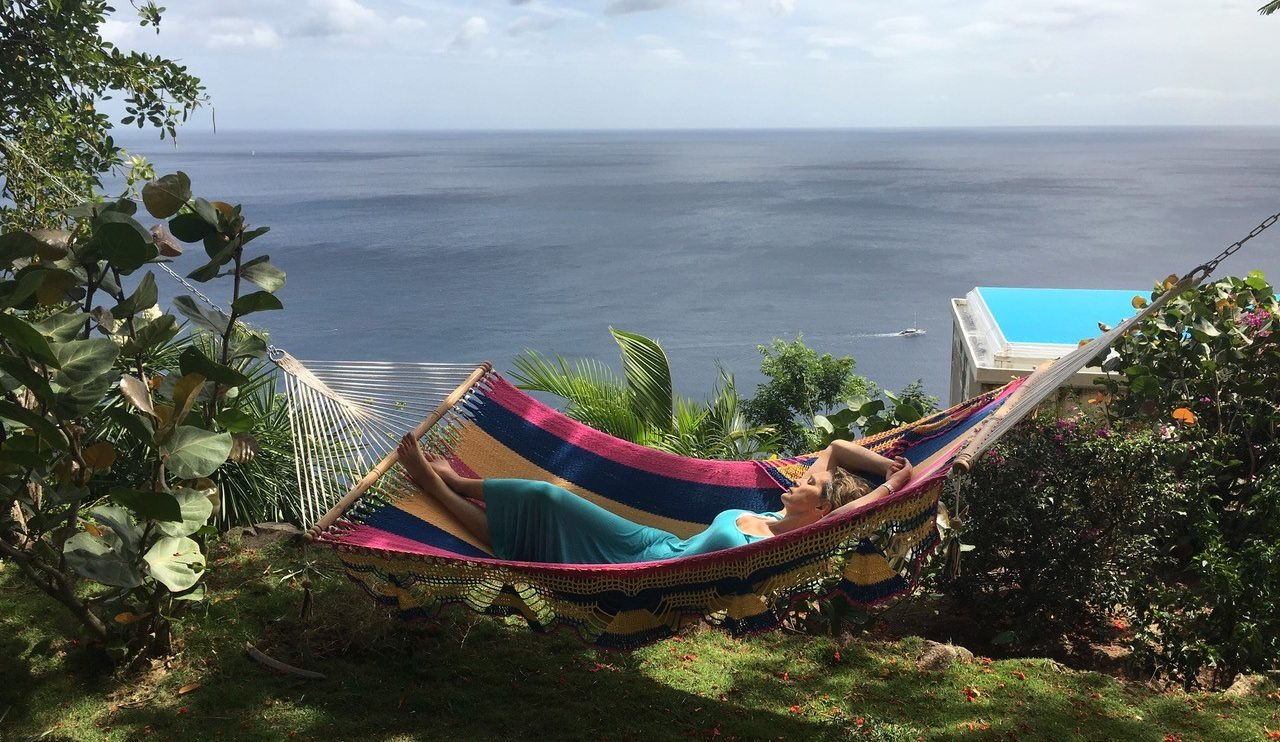 A woman relaxes in a colourful hammock at Cosmos St Lucia, with the infinity pool and Carribean Sea in the background