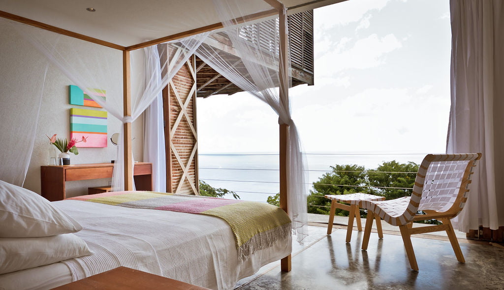 A four poster double bed, easy chair and desk in a Villa bedroom at Cosmos St Lucia, showing one wall open to the elements and the caribbean sea in the distance