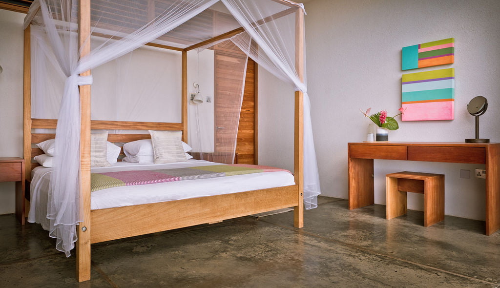 A four poster double bed in the Villa at Cosmos St Lucia, with matching wooden desk and stool, with bright coloured paiitnings on the wall and a gret concrete floor