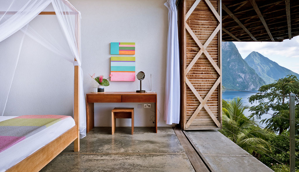 A double bedroom in the Villa at Cosmos St Lucia, featuring matching wooden desk and stool, bright paintings on the wall and a view of the Pitons