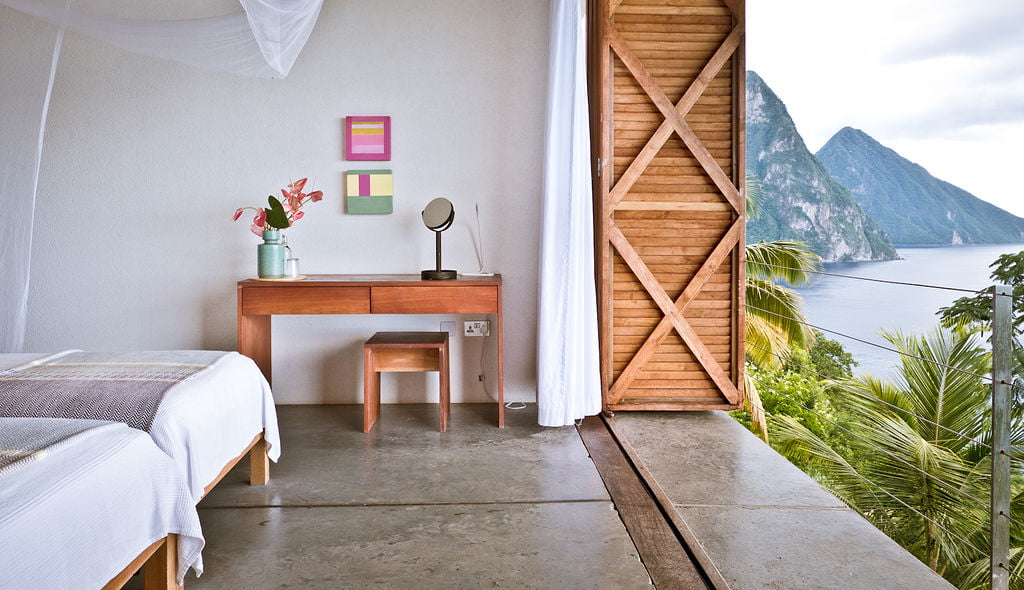 A twin bedroom at Cosmos St Lucia showing a desk and matching stool, paintings on the wall, wooden balcony doors and the Pitons in the distance