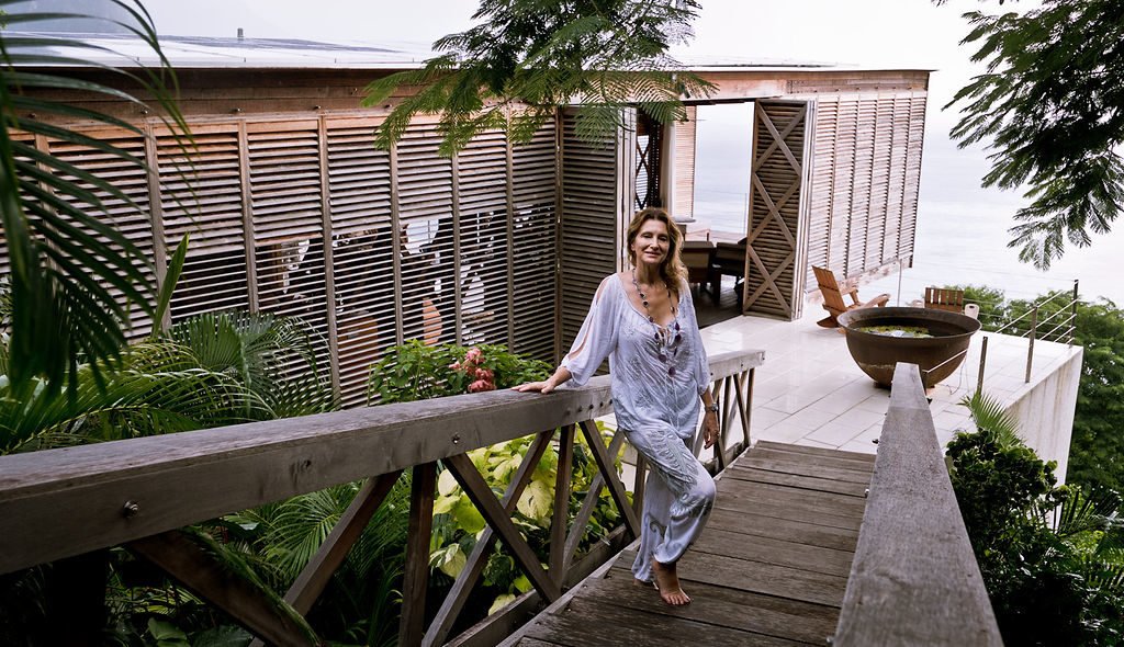 A woman wearing white stands on a wooden skybridge that leads to modernist wooden eco villa Cosmos St Lucia. The woman is smiling towards the camera
