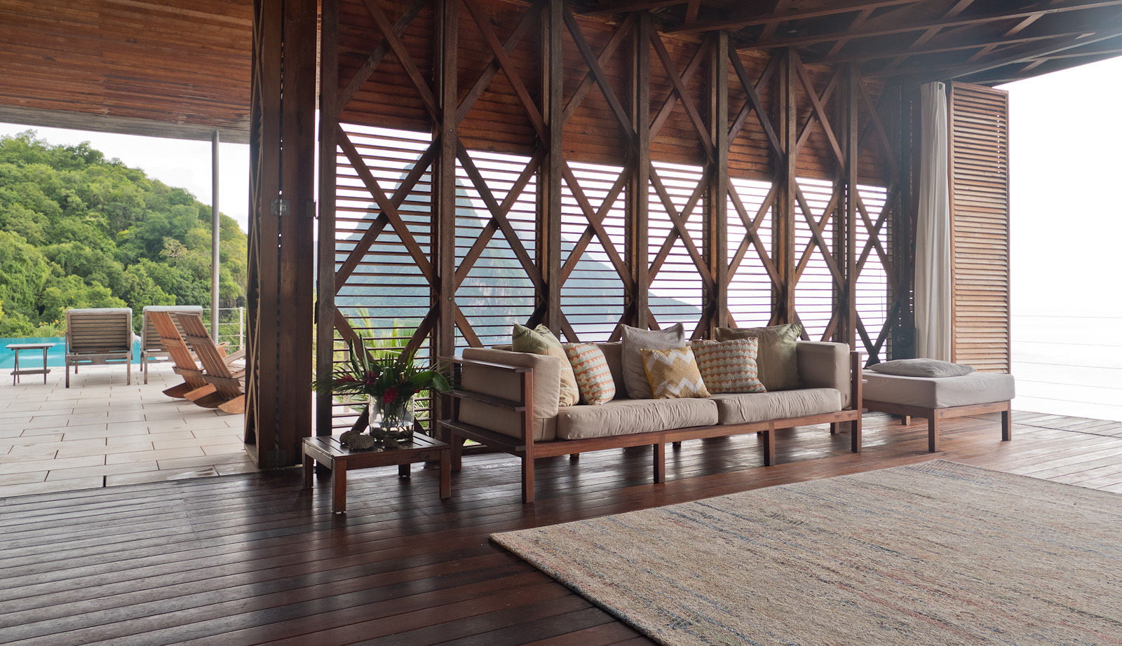 A view of the living space at Cosmos St Lucia, showing sofa seating, teak screens with concertina doors, and a sun terrace featuring sun loungers next to the pool. Rainforest hillside and the Pitons are visible beyond.