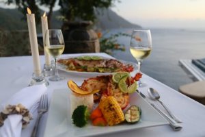 A dinner table set with a plate full of barbequed lobster and fresh vegetables, with a view of the Carribean sea in the background