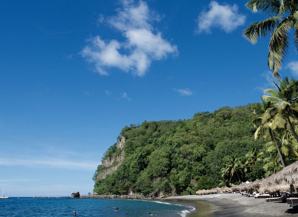 A blue sky above the cliffs covered in lush greenery next to the beach dotted with palm roofed parasols