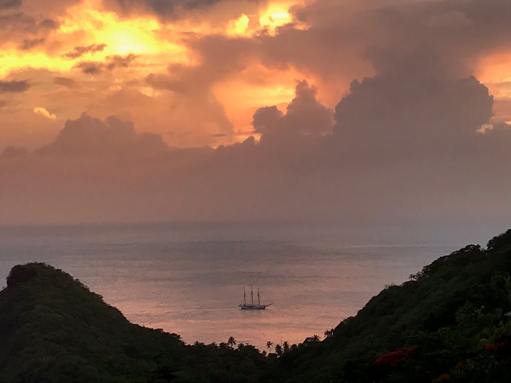 Pink sunset skies with the light of the sun breaking through, as the sun sets over the Caribbean Sea at Cosmos St Lucia. In the forground is the milly coastline and a three mast ship is visible on the sea