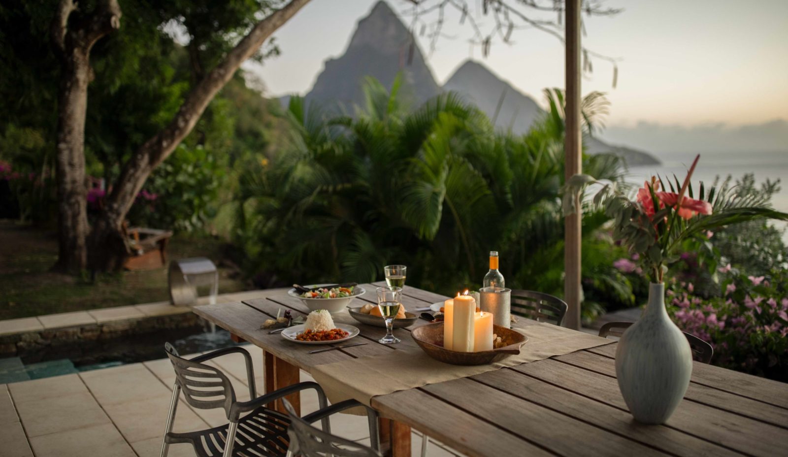 An outdoor wooden table holds an evening meal for two, with the garden at Cosmos St Lucia and the Pitons in the background