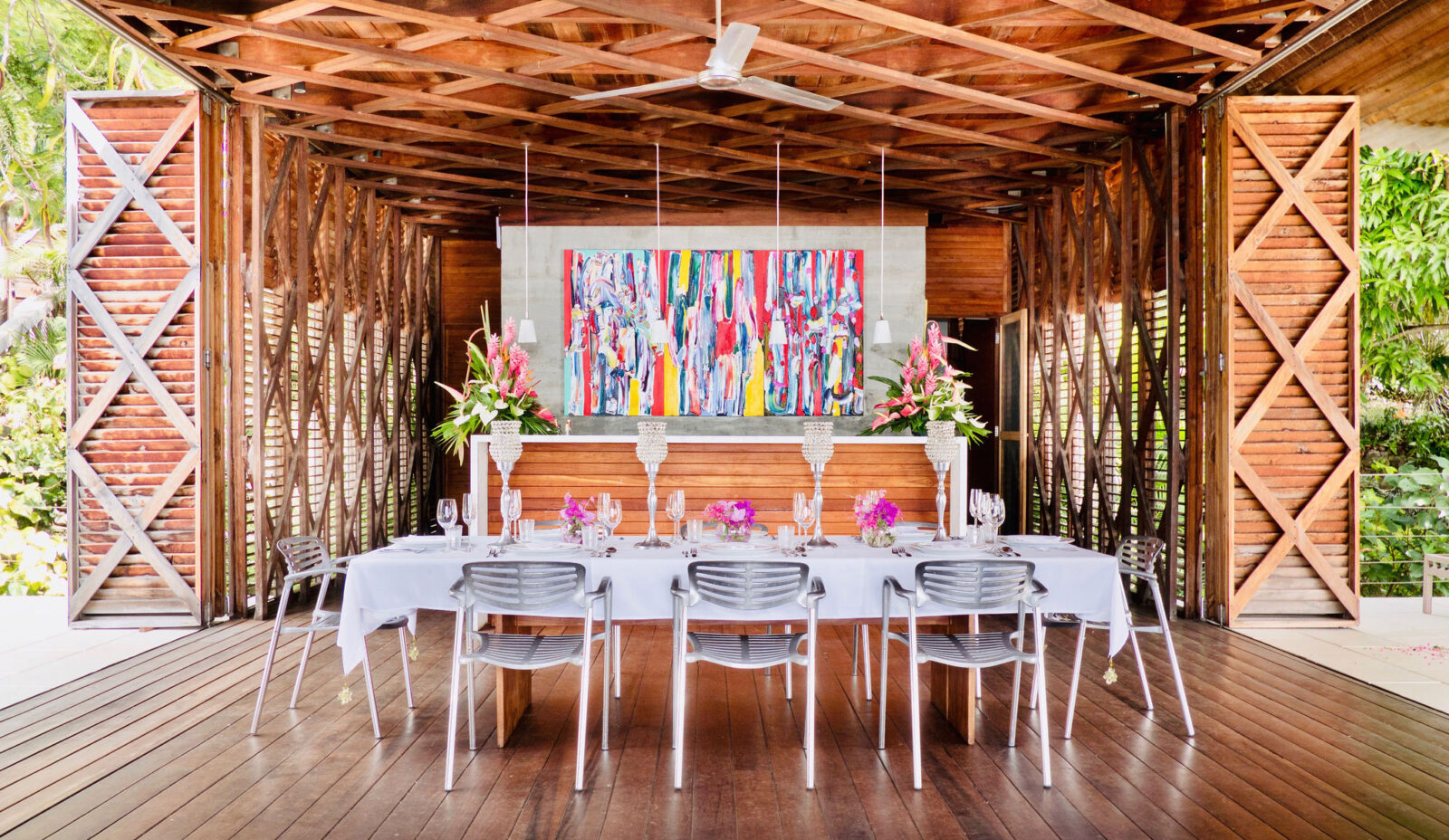 A celebratory table setting for 8 people featuring white and silver tableware, large floral arrangements and a brightly coloured painting in an entirely wooden Cantilever at Cosmos St Lucia