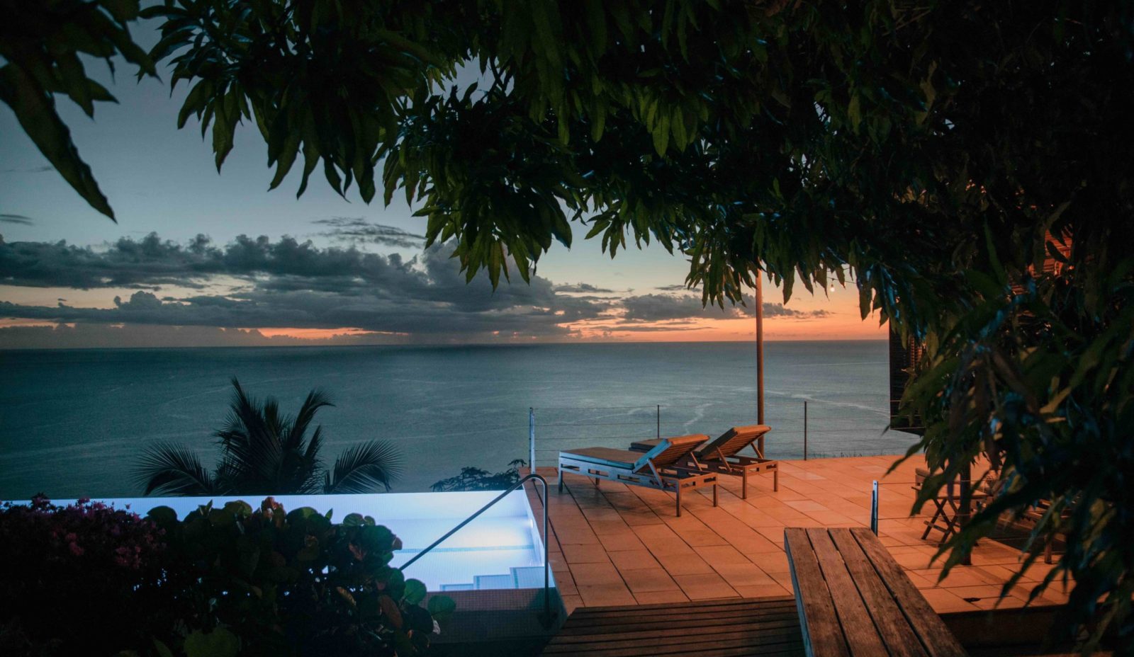 Dusk on the sunterrace at Cosmos St Lucia, featuring the pool and sun loungers framed by foliage in the foreground, with sunset over the Caribbean Sea in the background