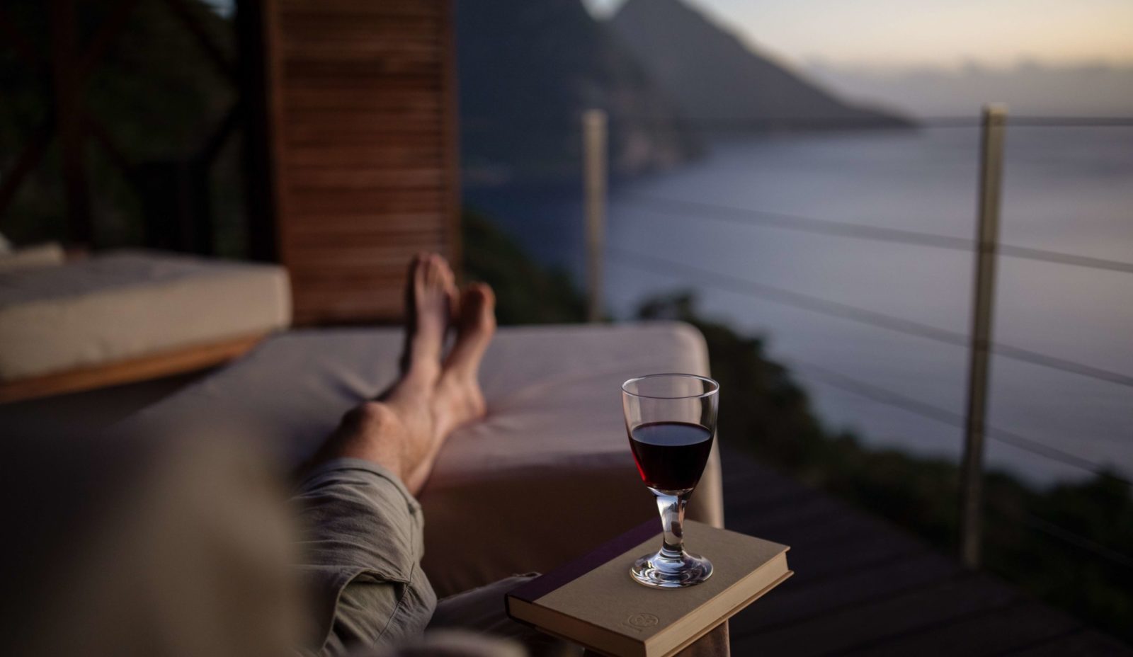 A man is reclining in a soft arm chair while looking at the Pitons from the Cantilever at Cosmos St Lucia. His legs are stretched out and crossed and there's a book and a glass of red wine by his side