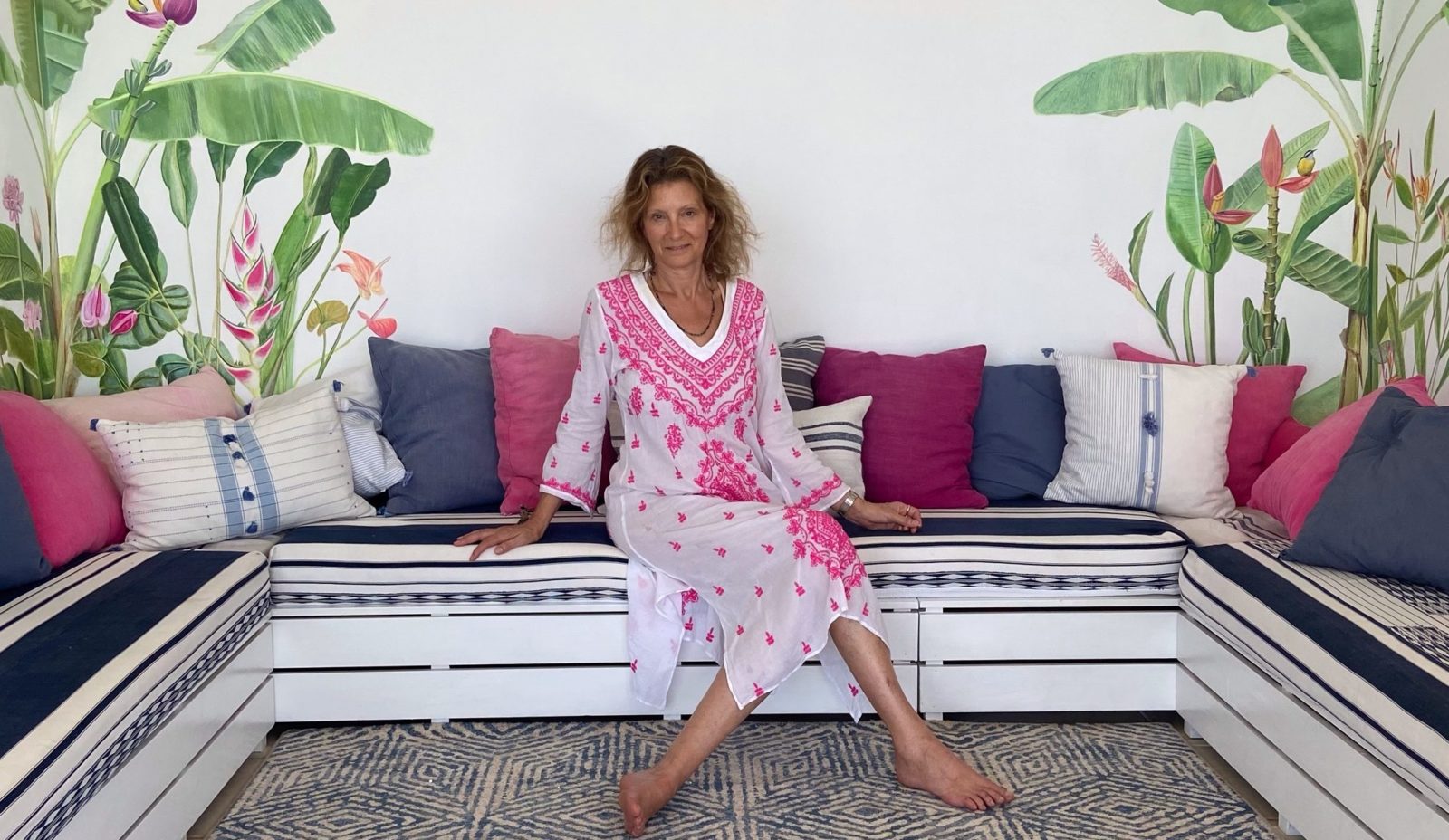 A woman in a white and pink dress sits on striped cushions in a white room decorated with colourful murals inspired by the flora and fauna in the surrounding rainforest at Cosmos St Lucia