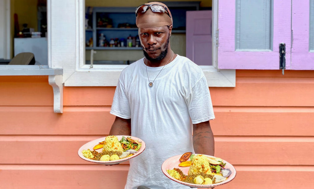 A St Lucian man stands in front of an orange and pink painted shack, holding two plates of food he has prepared to order for customers. he wears a white T shirt and blue shorts and stairs into the camera