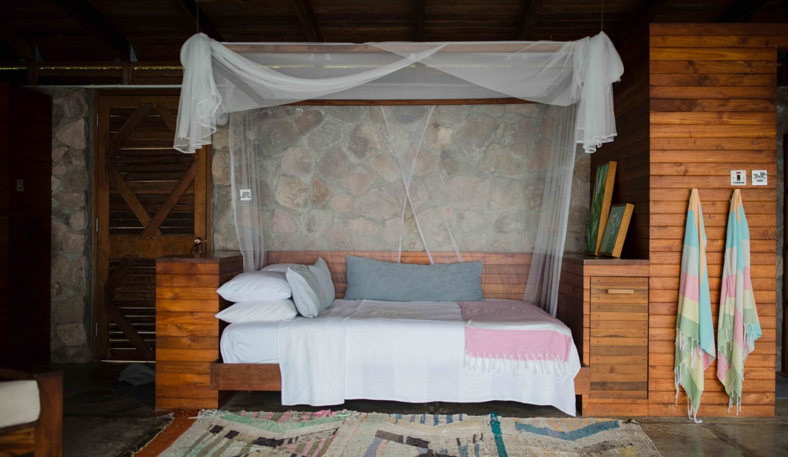 The queen size bed in the Lodge at Cosmos St Lucia has a wooden frame above it holding a mosquito net. There's a pale patterned rug on the floor and two coloured turkich towels hang on the wall. The background shows the stone outwall and wooden pannelling inside