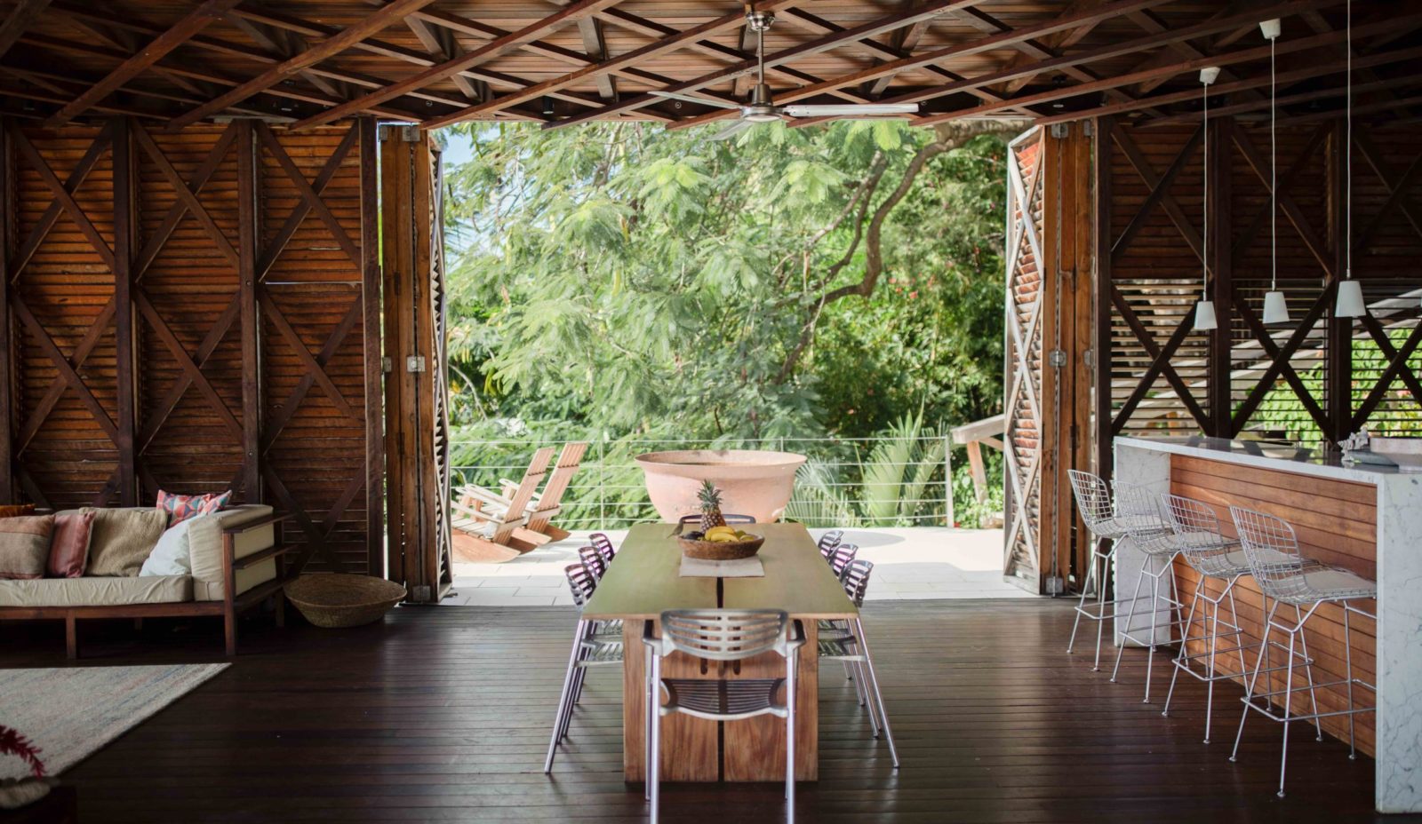 The dining area inside the wooden Cantilever at Cosmos St Lucia features a dining table for 8, with the Marble Bars and 4 stools on the right, and lounge seating on the left, looking out onto a mango tree in the garden