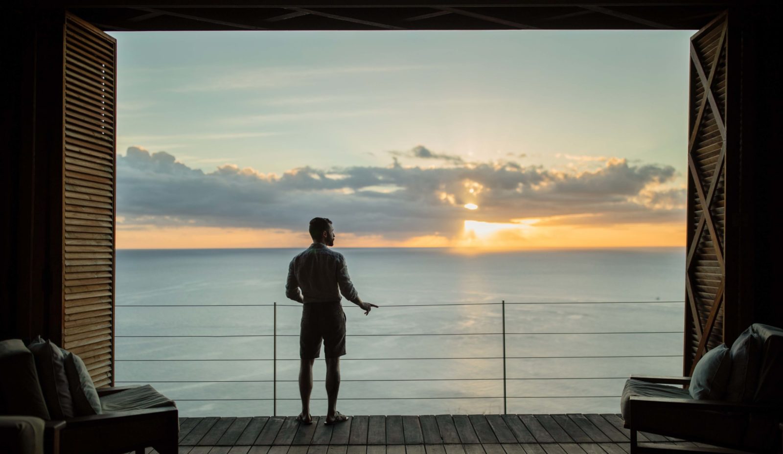 A man stands looking out over the sunset in the Cantilever at Cosmos St Lucia. Seating features on either side of the foreground, with the sunset in the distance