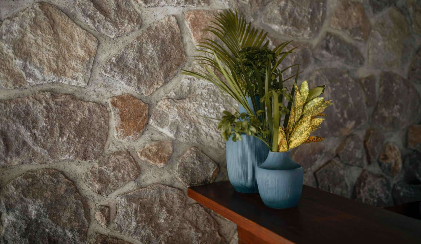 Two blue vases are filled with yellow and green foliage from the garden at Cosmos St Lucia, shown sitting on a wooden surface and against a stone wall