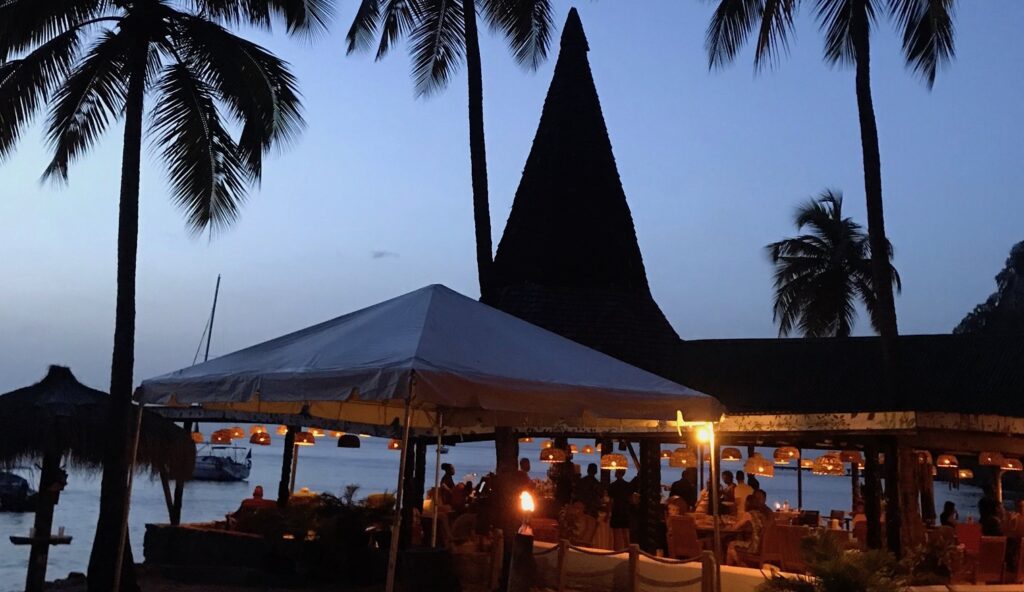 An inviting, open air beach bar at dusk, with soft lighting hanging from the ceiling and the silouhettes of palm trees overhead, with the sea and boats beyond