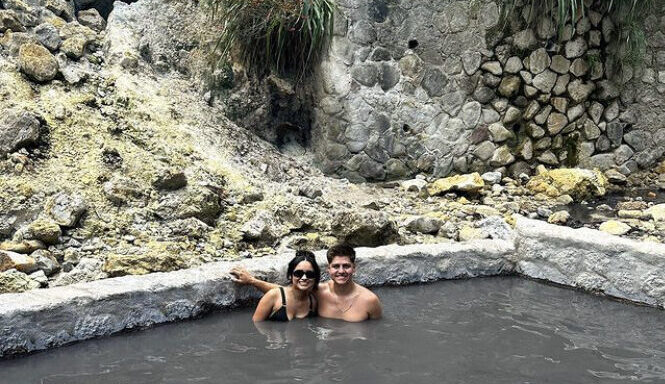 A couple in their swimwear relax in a mudbath in a volcanic landscape of grey rock and scrub