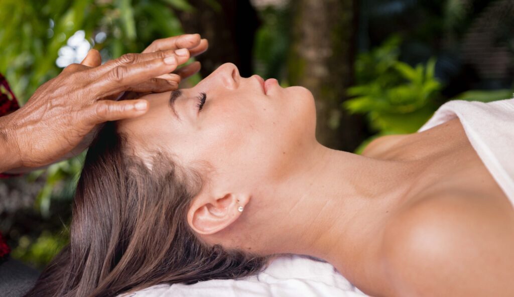 The hands of a massage therapist gently massage the brow of a woman lying with her eyes closed on a massage table. Green leaves and trees are visible in the background