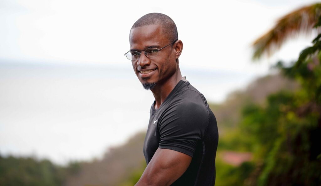 A brown skinned man with close cropped hair smiles into the camera. He wears rimless glasses and a short goatee. His form fitting black T shirt shows his muscular physique, befitting for a personal trainer