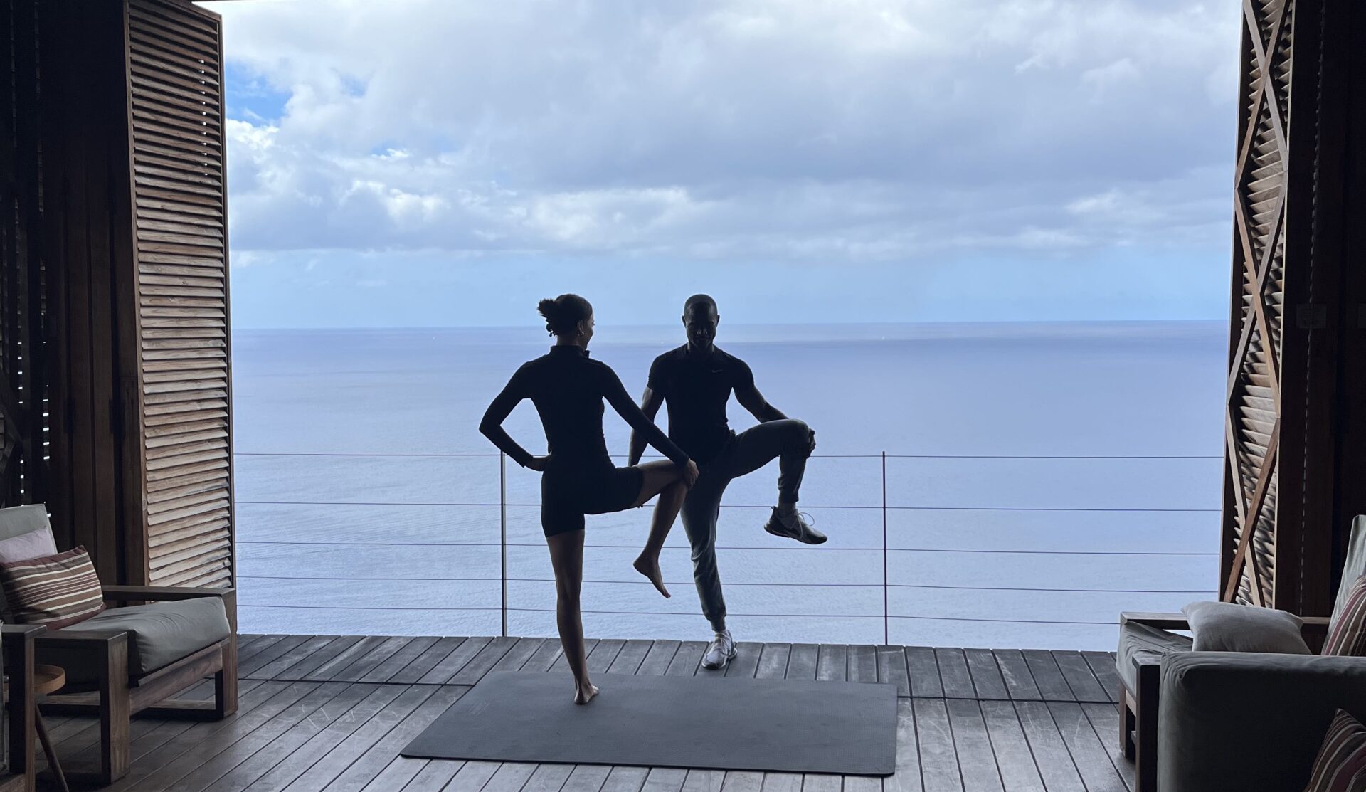 Two people wearing gym clothes face each other, bending and stretching one leg to the side. Behind them is the sea