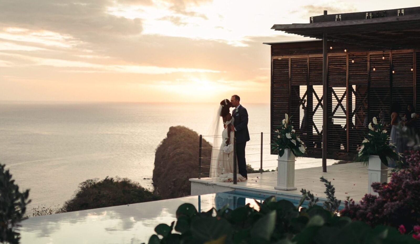 An interacial wed couple enjoy a quiet embrace ona terrace with the sea and sunset behind them