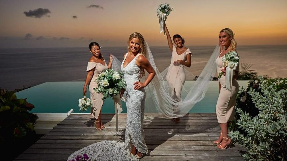 A bride is flanked by 3 bridesmaids holding her veil and flowers, standing in front of a sunset sea and sky