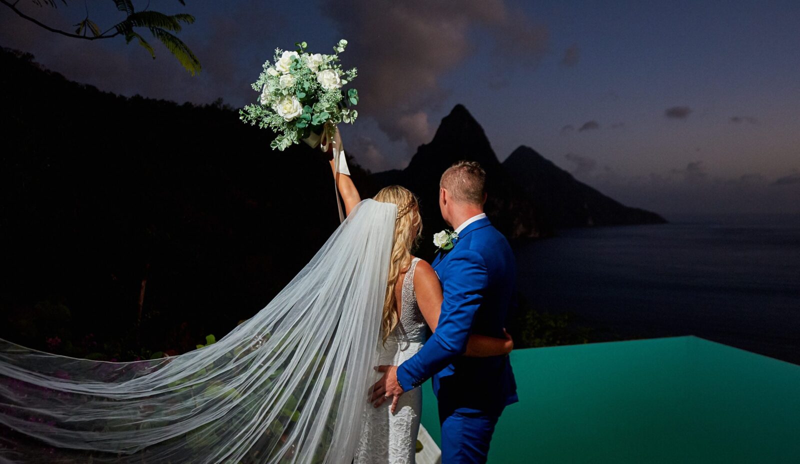 A newly wed couple face away from the camera as the bride prepares to throuw her bouquet. They stand infront of a turquoise pool and the pitons mountains in the distance