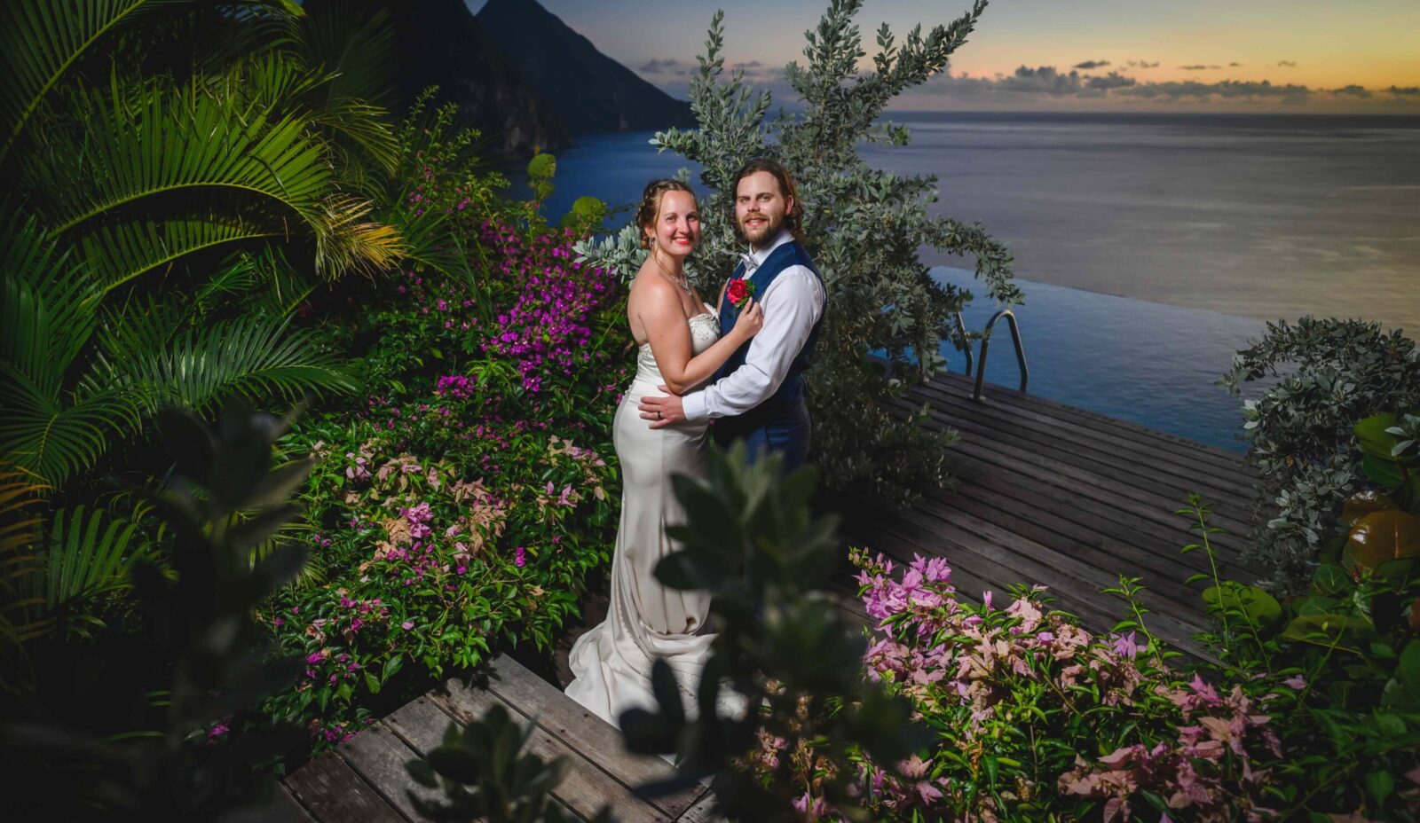 A just married couple smile at the camera, with flowers and greenery on either side, the sea and pitons mountains behind them