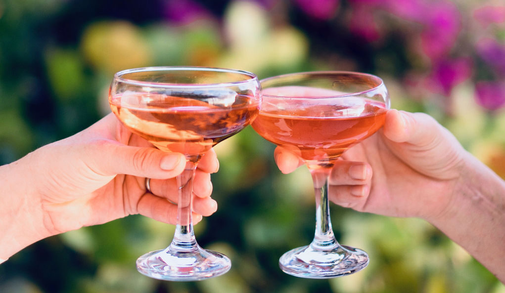 Two coupe glasses of pink champagne are chinking together in a cheers gesture, each held by a single hand of a white person, with garden greenery blurred behind them